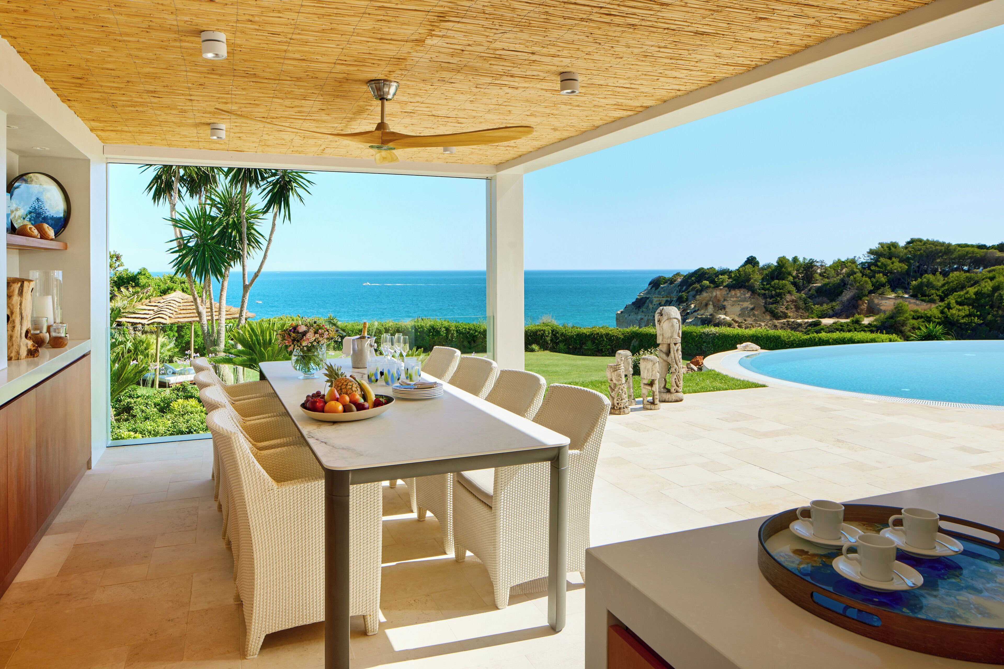 A dining area with a view of the ocean, a pool, and a patio with chairs and a table.