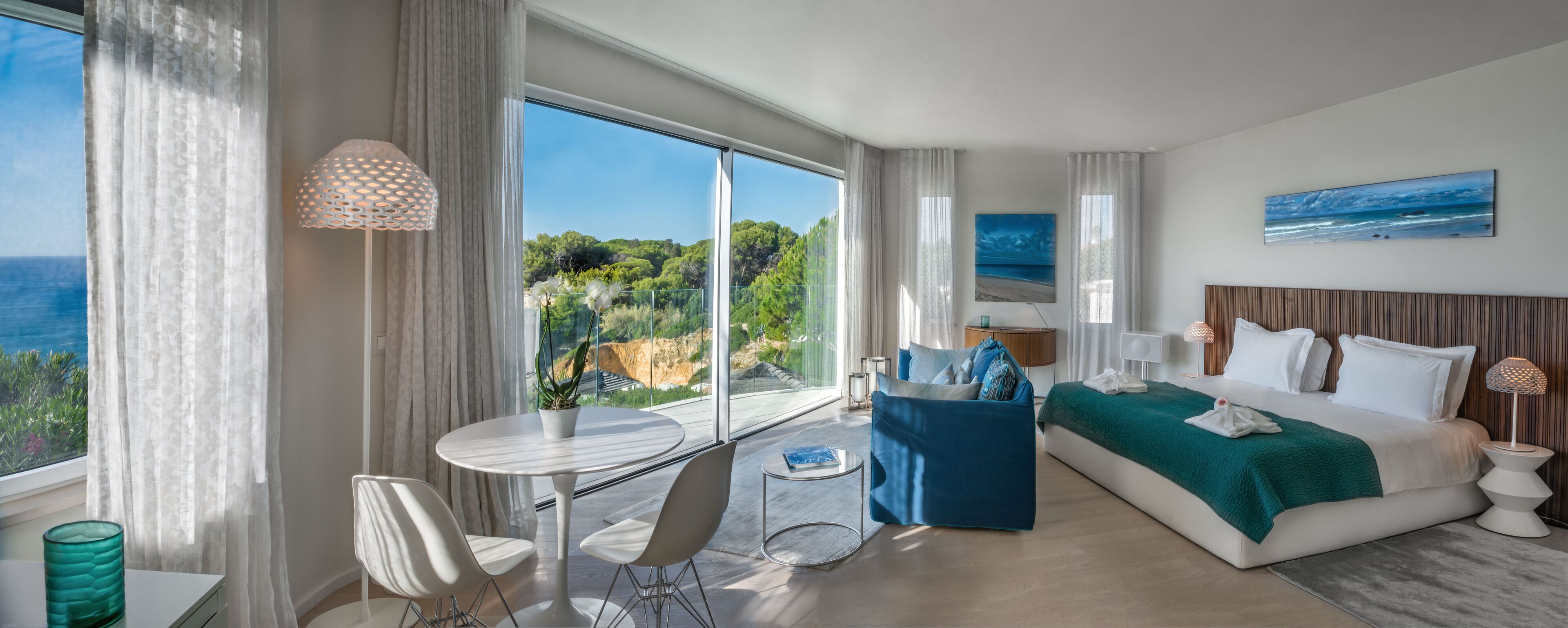 Habitación de hotel moderna con un balcón grande, una cama con una manta verde, una mesa de comedor redonda y vista al mar.