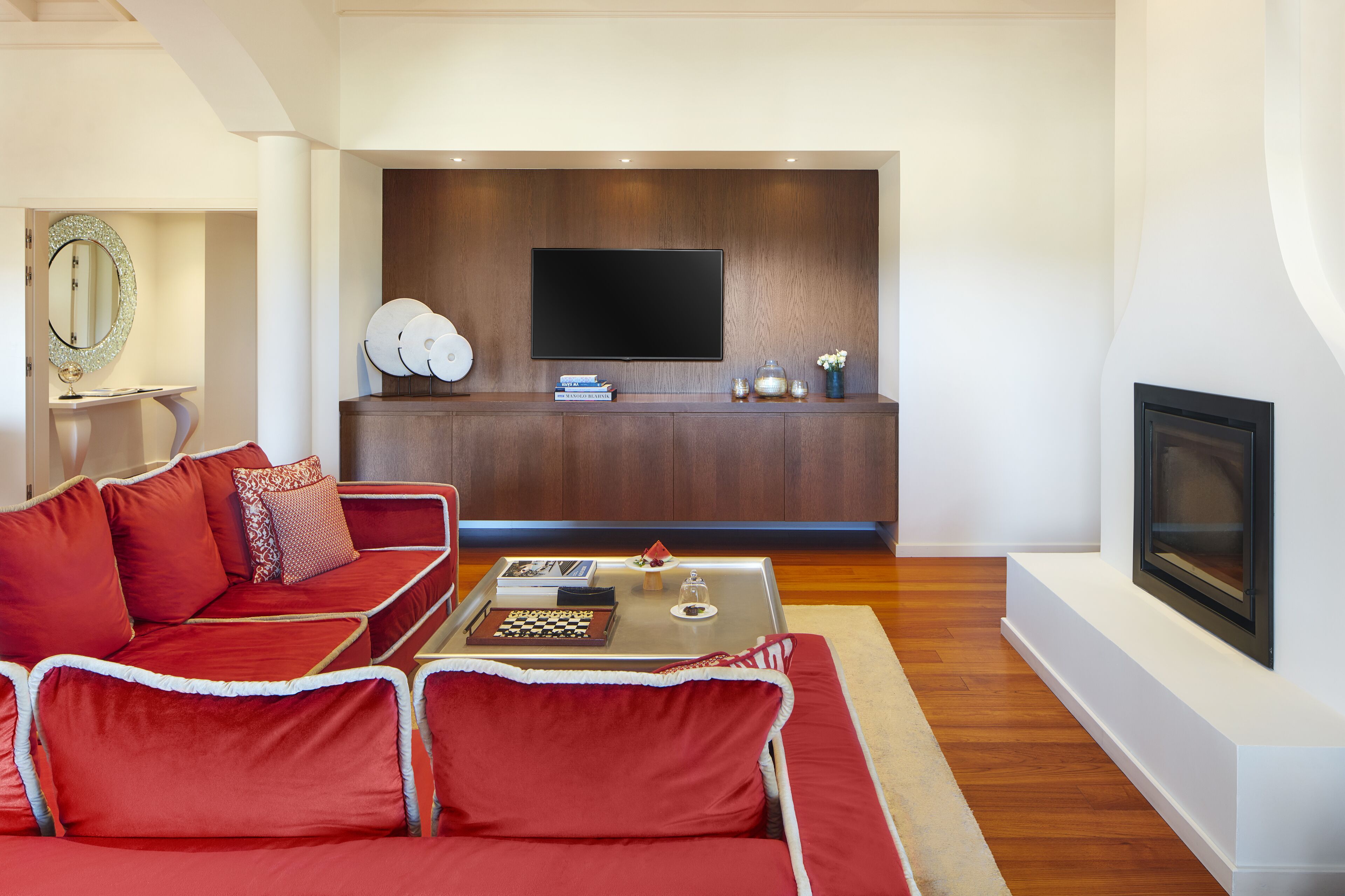 A modern living room with a red sofa, a coffee table with books and a chessboard, a flat-screen TV mounted on a wooden wall, and a fireplace.