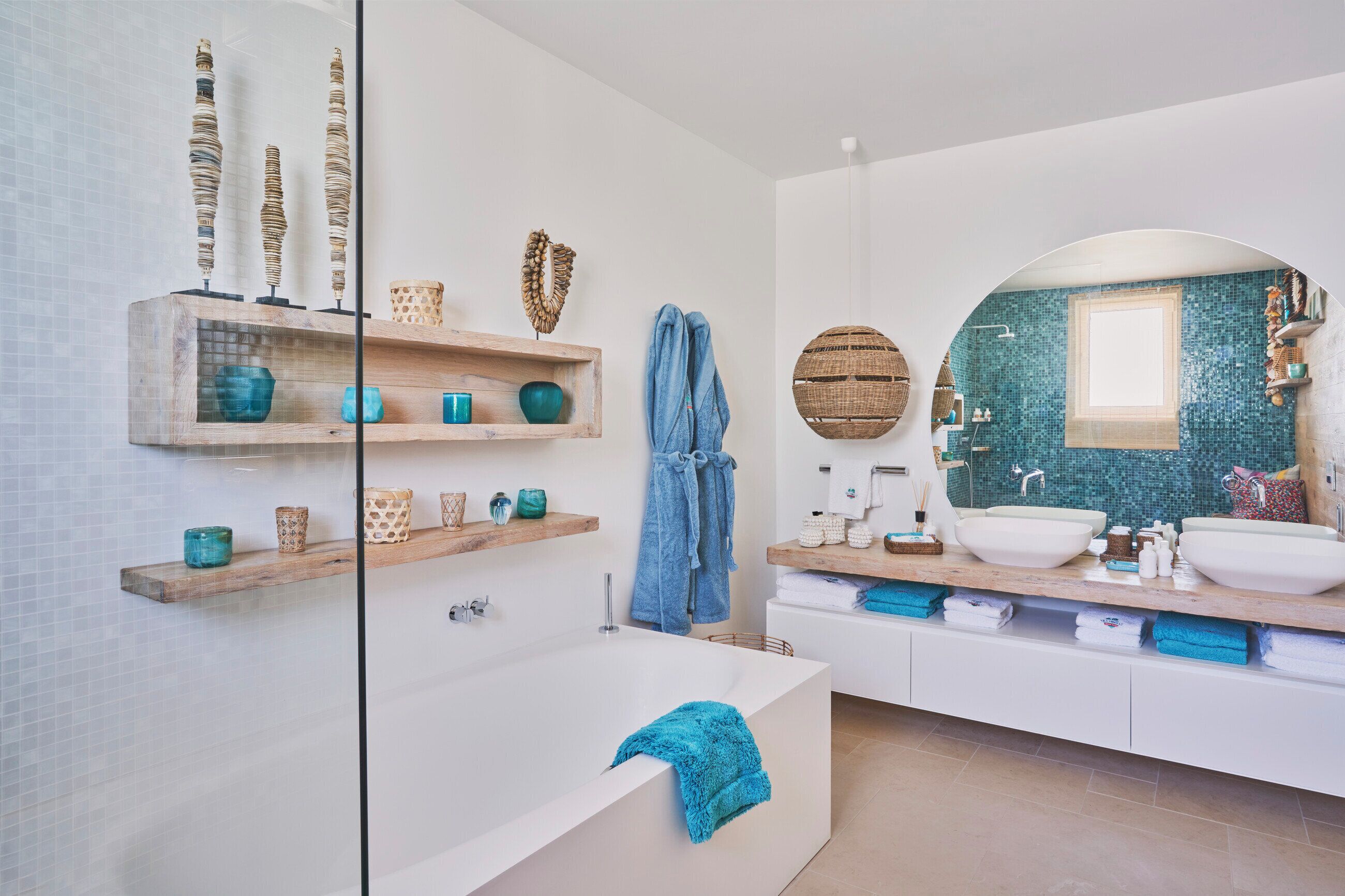 Un baño moderno con una bañera grande, un lavamanos doble y un espejo amplio. Se observan estantes de madera con objetos decorativos y un perchero para toallas.
