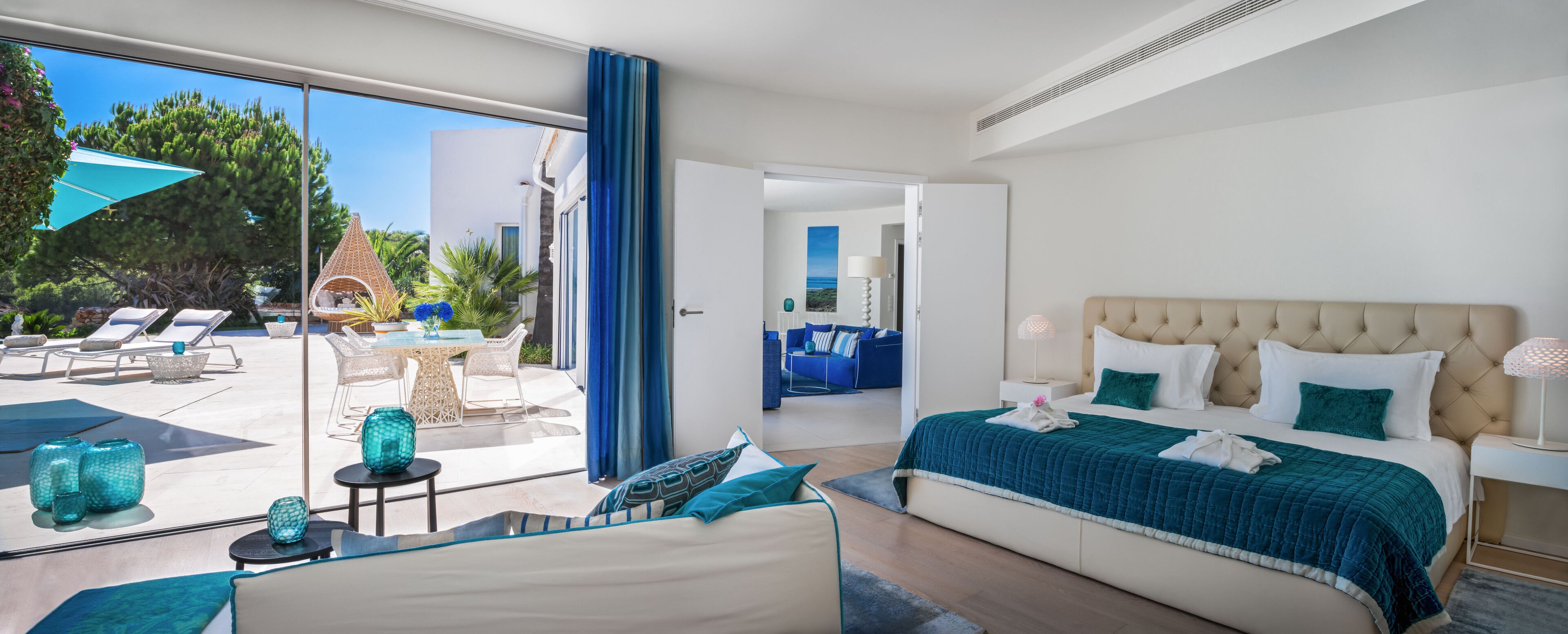 A modern hotel room with a large bed, a balcony with lounge chairs, and a view of the outdoors.