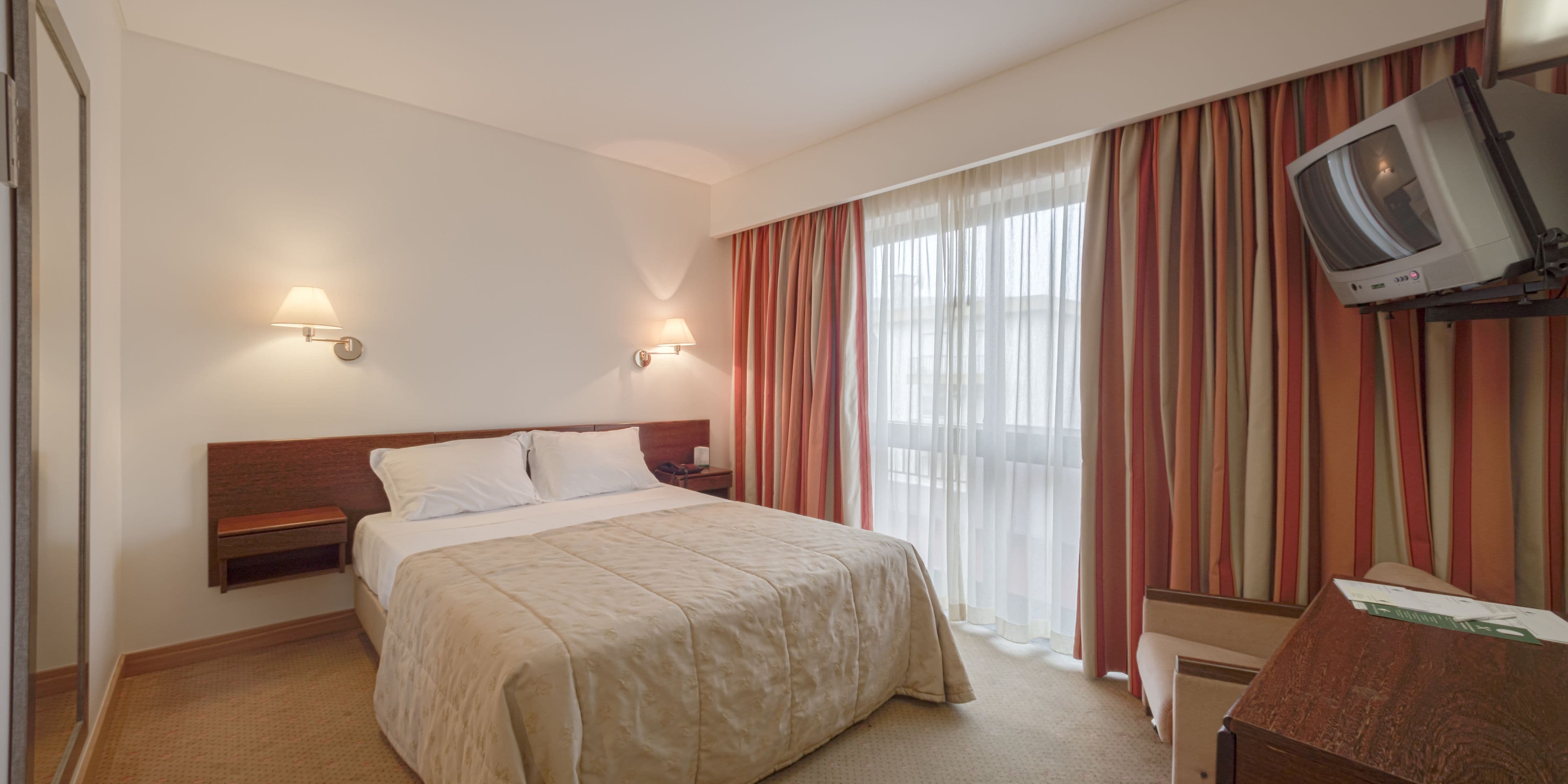A hotel room with a bed, bedside tables, a TV mounted on the wall, and curtains.