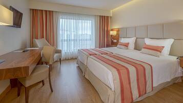 Quarto de hotel com cama, escrivaninha, cadeira, televisão e janela com cortinas.