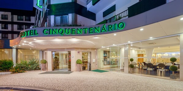 Hotel Cinquentenário features a modern entrance with a curved overhang and glass facade, flanked by potted plants and a cobblestone pathway.