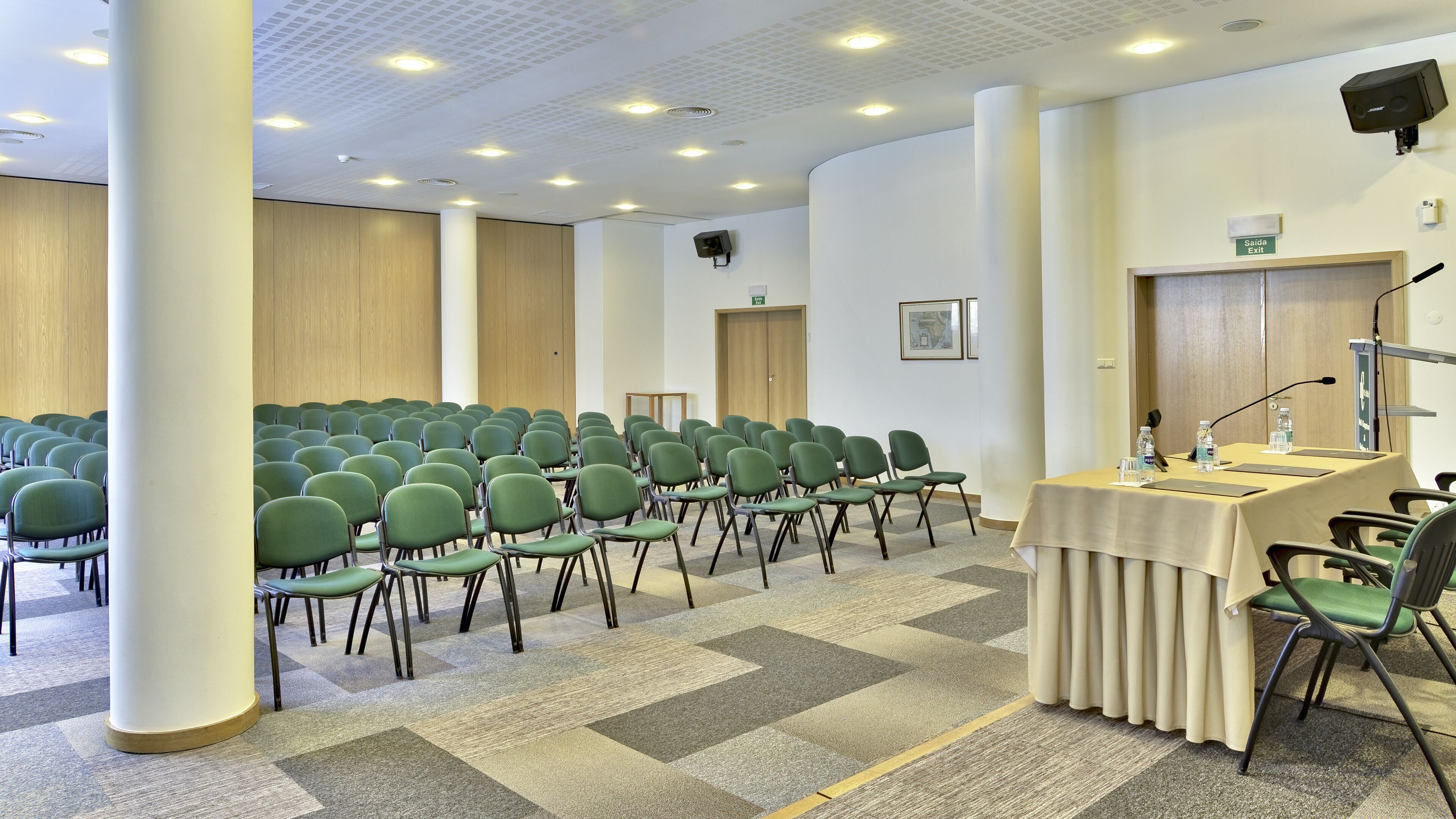 A conference room with rows of green chairs, a podium with a microphone, and a "Saída Exit" sign.