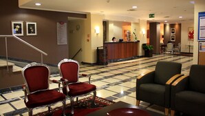 A hotel lobby with a reception desk, two red upholstered chairs, a green chair, and a patterned carpet. - Hotel Orquidea (Funchal)