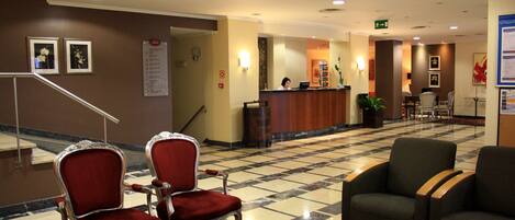 A hotel lobby with a reception desk, two red upholstered chairs, a green chair, and a patterned carpet.