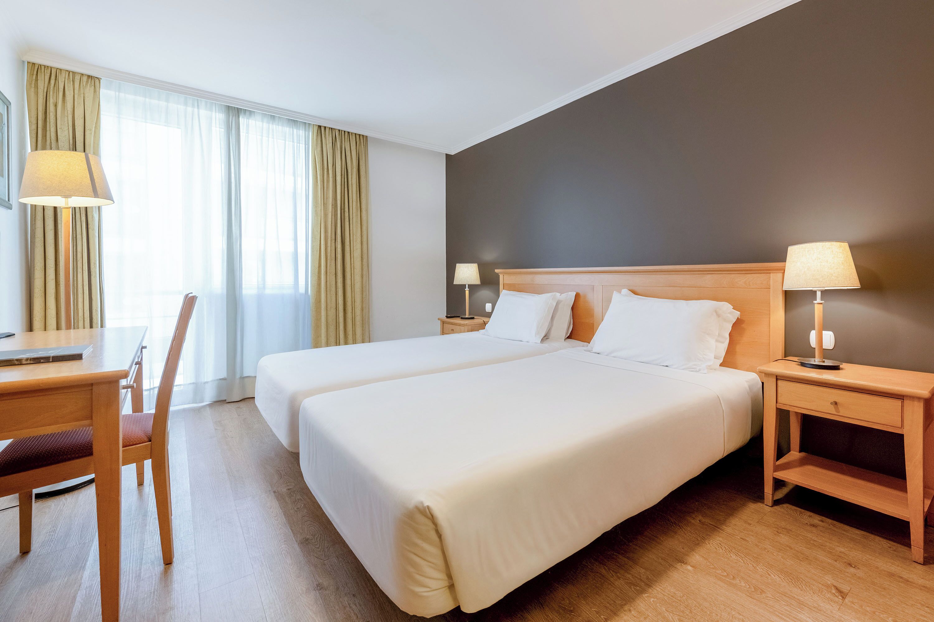 A hotel room with two beds, a desk, a chair, and a nightstand with lamps.