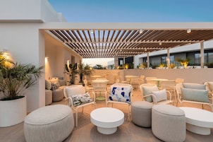 Un espace extérieur aménagé avec des tables rondes et des chaises en osier blanc, entouré de plantes en pot et d’une pergola en bois.