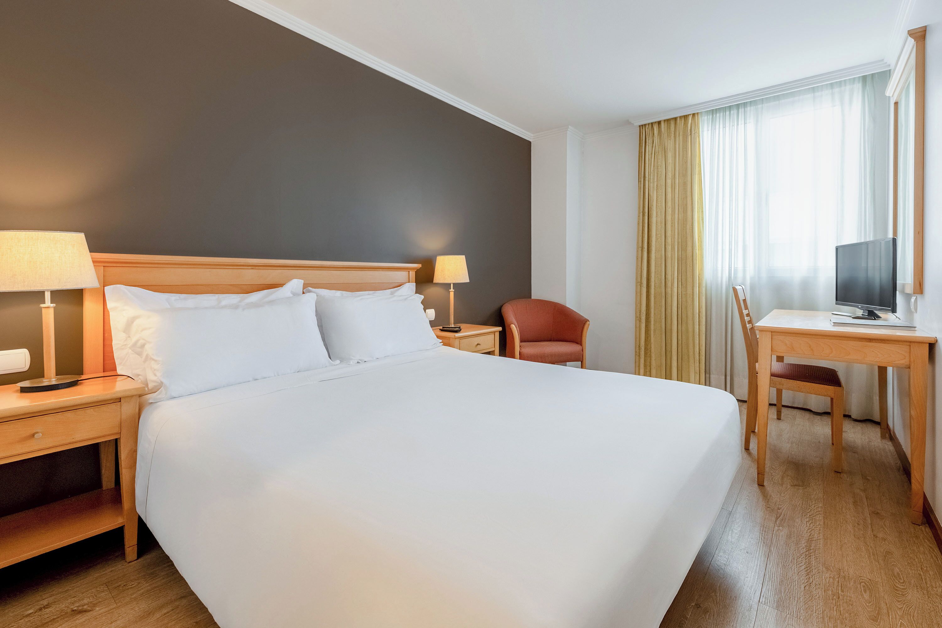 A hotel room with a large bed, a desk, a chair, a TV, and a lamp.