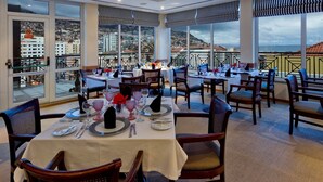 A restaurant with tables set for dining, large windows offering a city view, and a balcony with a railing. - Hotel Orquidea (Funchal)