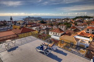 Una terraza en la azotea con vistas a un pueblo costero, que presenta una mezcla de arquitectura tradicional y moderna, y un crucero en la distancia.