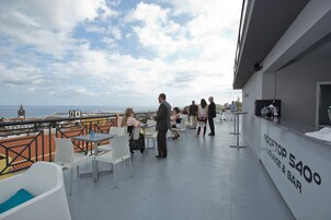 A rooftop lounge area with tables and chairs, a bar, and a view of the city and sea.