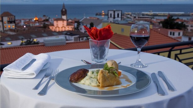 A table with a meal, a glass of red wine, and a glass of water, overlooking a coastal cityscape. - Hotel Orquidea (Funchal)