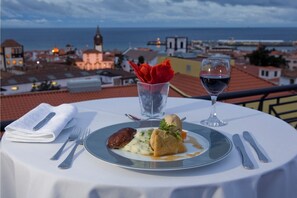 A table with a meal, a glass of red wine, and a glass of water, overlooking a coastal cityscape. - Hotel Orquidea (Funchal)