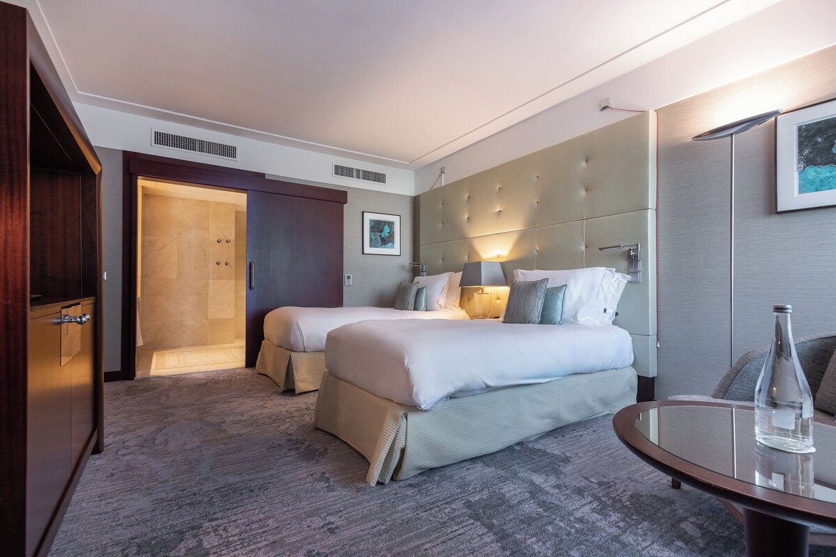 Habitación de hotel con dos camas, una zona de estar con mesa y un espacio para la ducha.