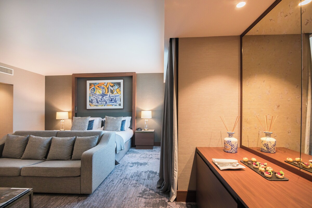 A hotel room with a bed, a sofa, a bedside table with lamps, a mirror, and a tray of snacks.