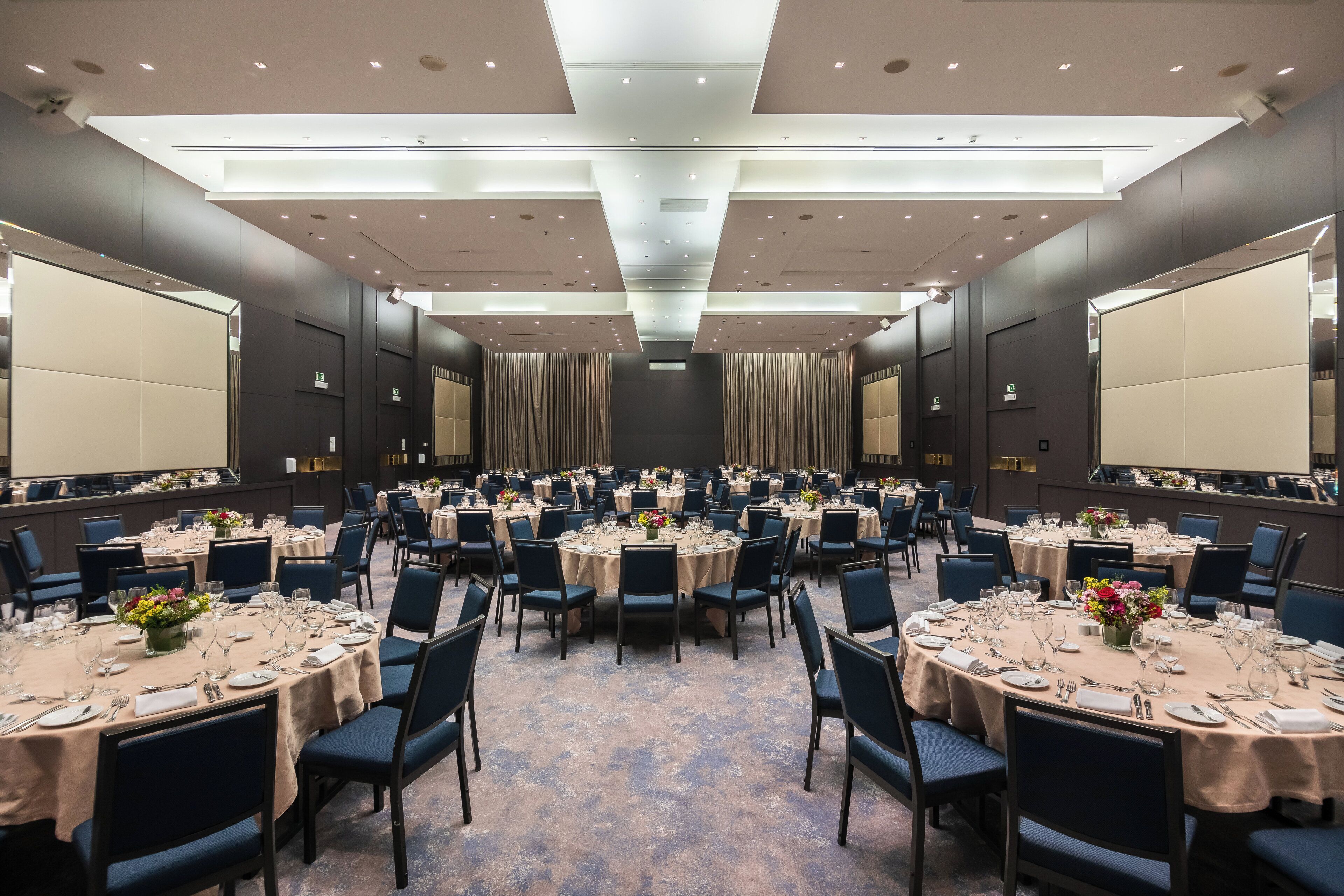 A large conference room with multiple round tables, each set with chairs, plates, and cutlery.