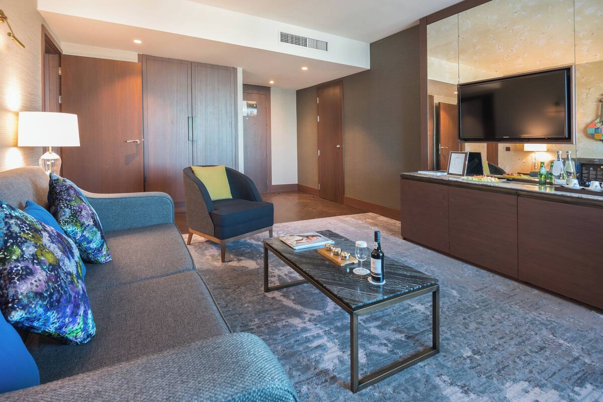 A modern living room with a sofa, armchair, coffee table, and a flat-screen TV mounted above a kitchen counter.