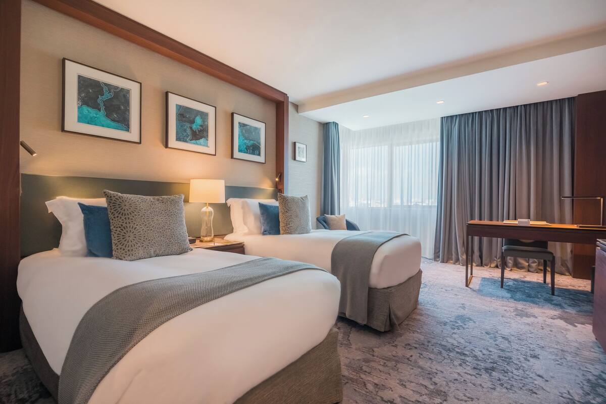 A hotel room with two beds, a desk, and a large window with curtains.