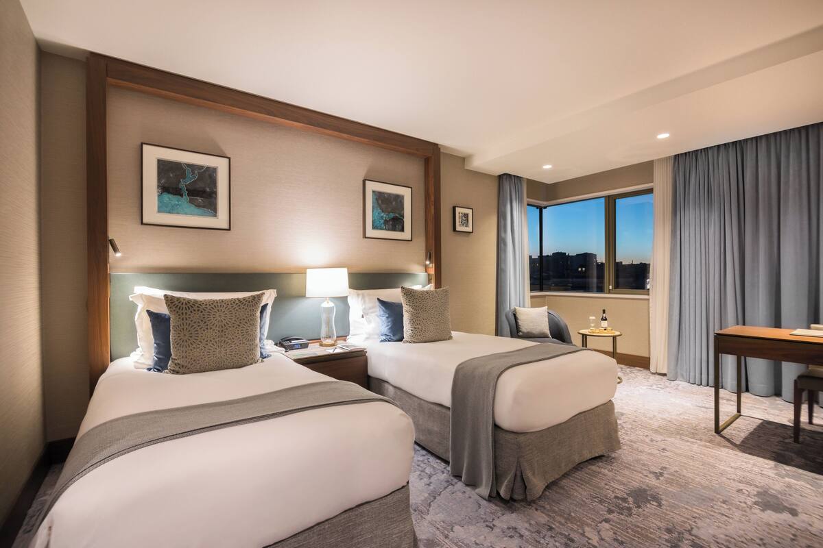 A hotel room with two beds, a desk, a chair, and a window with curtains.