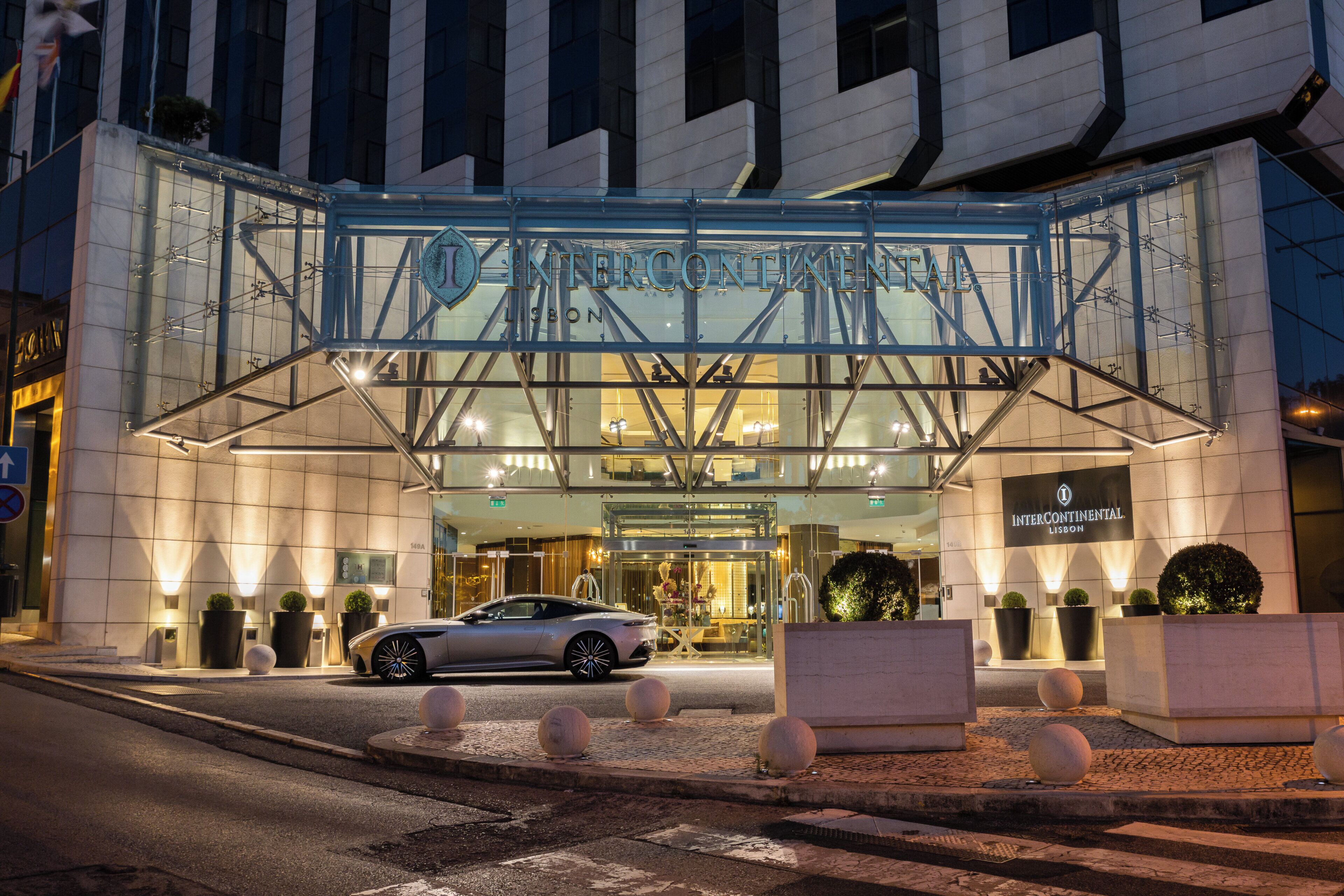 The Intercontinental Lisbon hotel entrance with a modern glass facade, a car parked in front, and a well-lit interior visible through the glass.