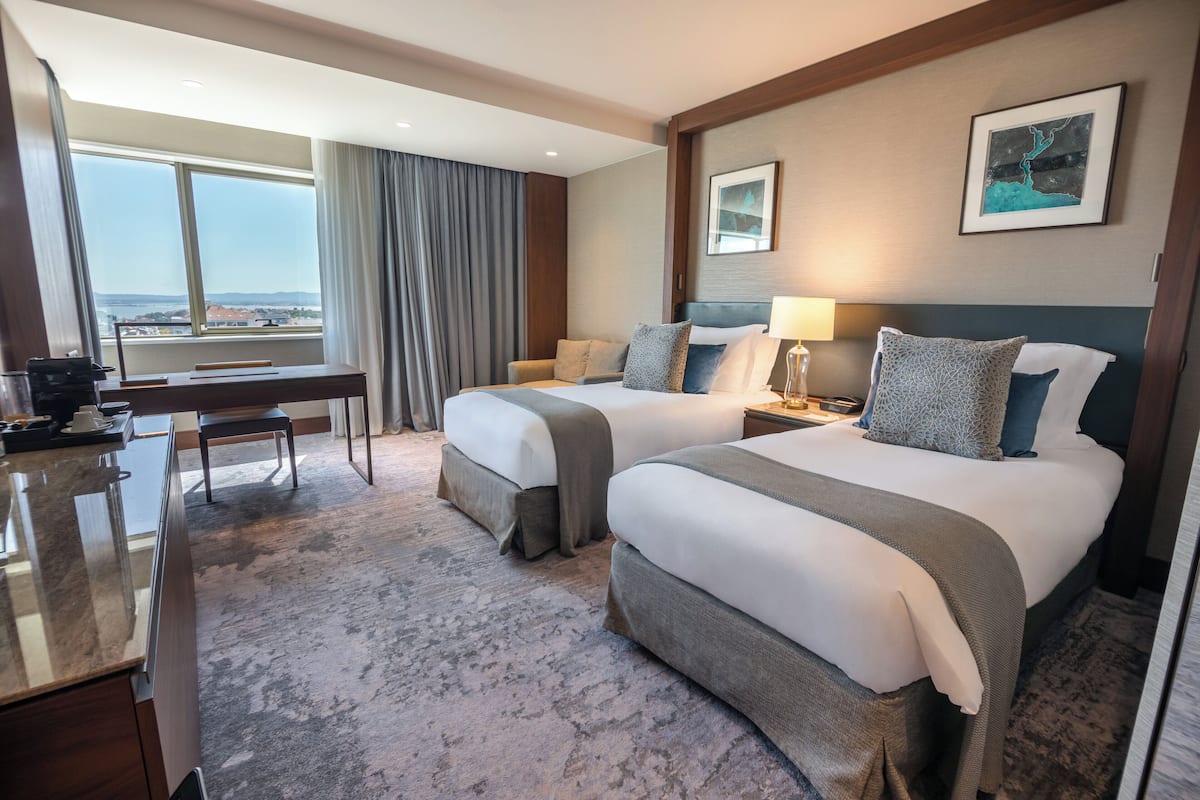 A hotel room with two beds, a desk, and a large window offering a city view.