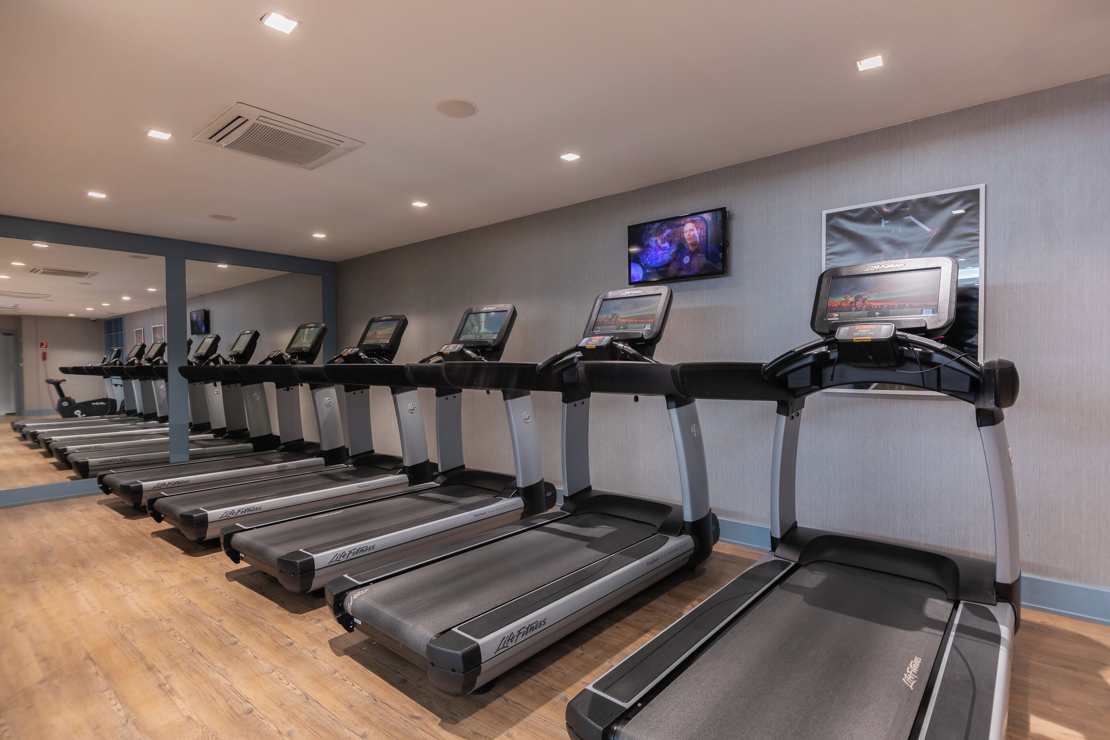 A row of treadmills in a gym with a TV on the wall and a mirror reflecting the space.