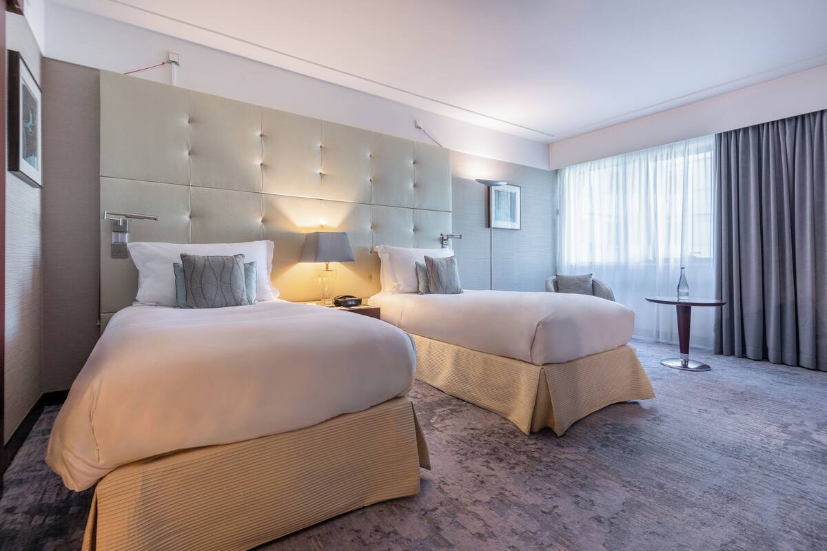 A hotel room with two beds, a large padded headboard, a small table, and a view of the outdoors through large windows.