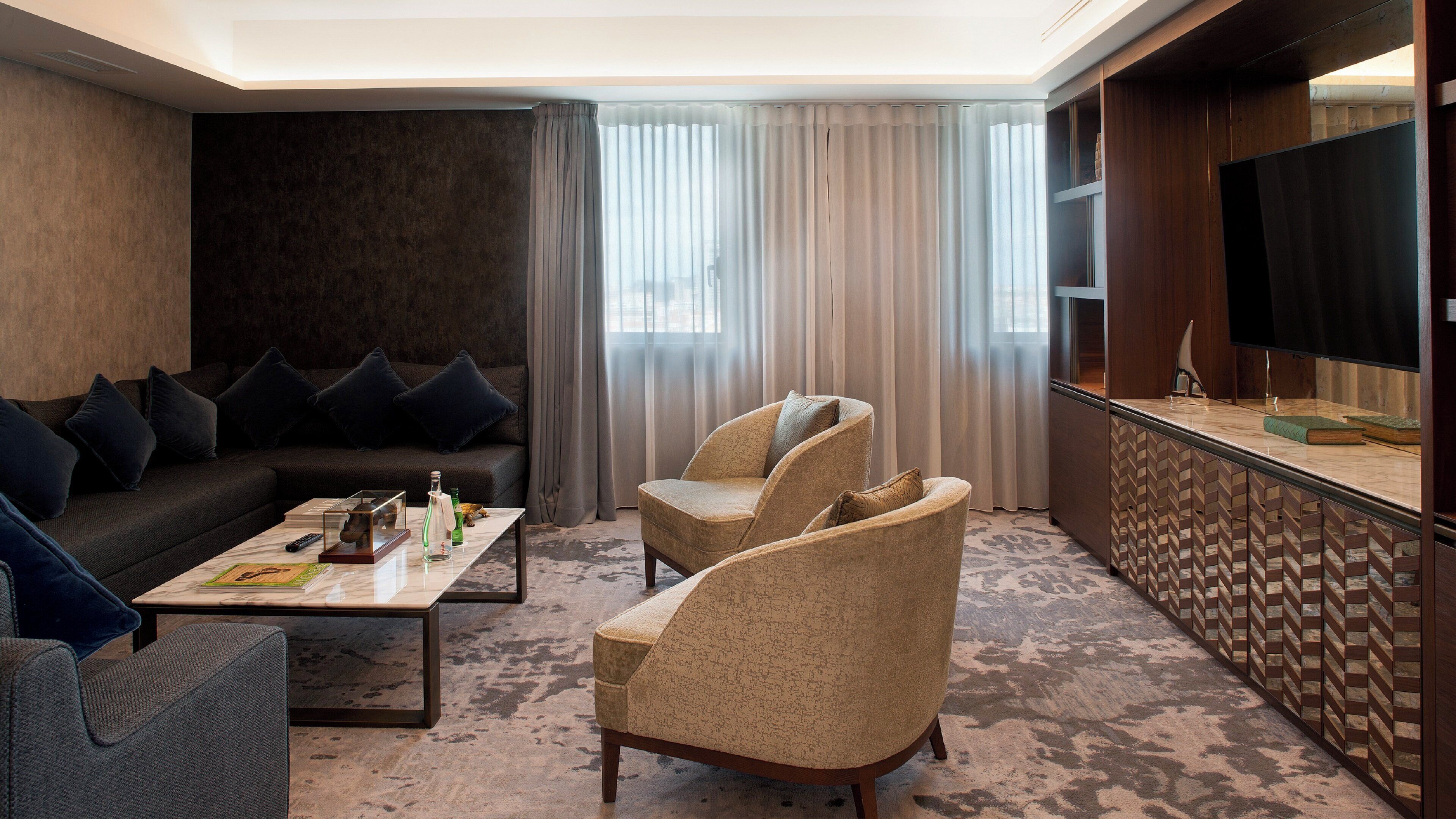A modern hotel room with a dark-colored sofa, two armchairs, a coffee table, and a flat-screen TV.