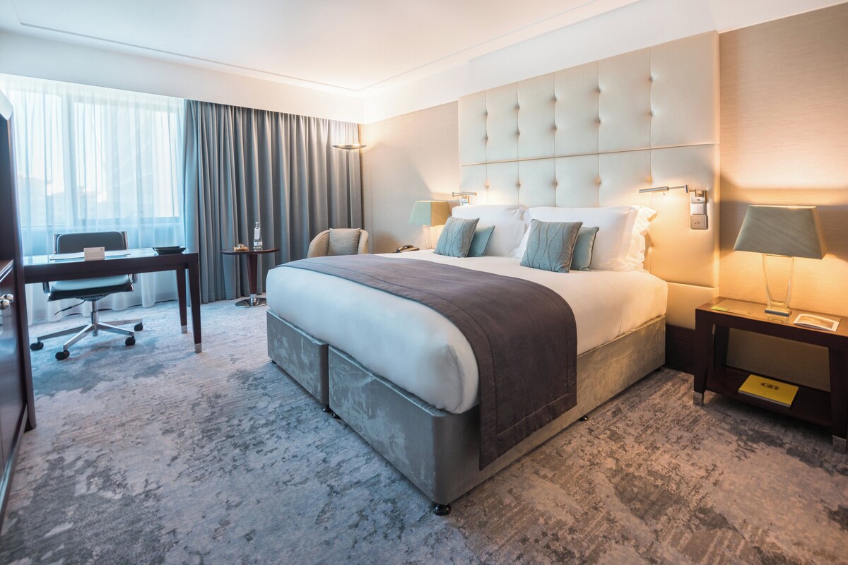 A hotel room with a large bed, a desk, a chair, and a view of the outdoors.