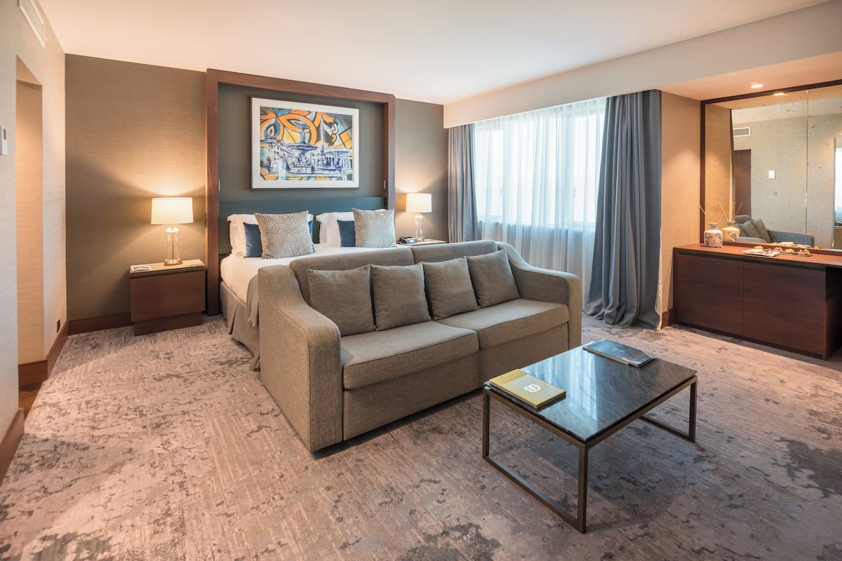 A modern hotel room with a sofa, a coffee table, a bed with pillows, a nightstand with a lamp, a painting on the wall, and a large mirror.