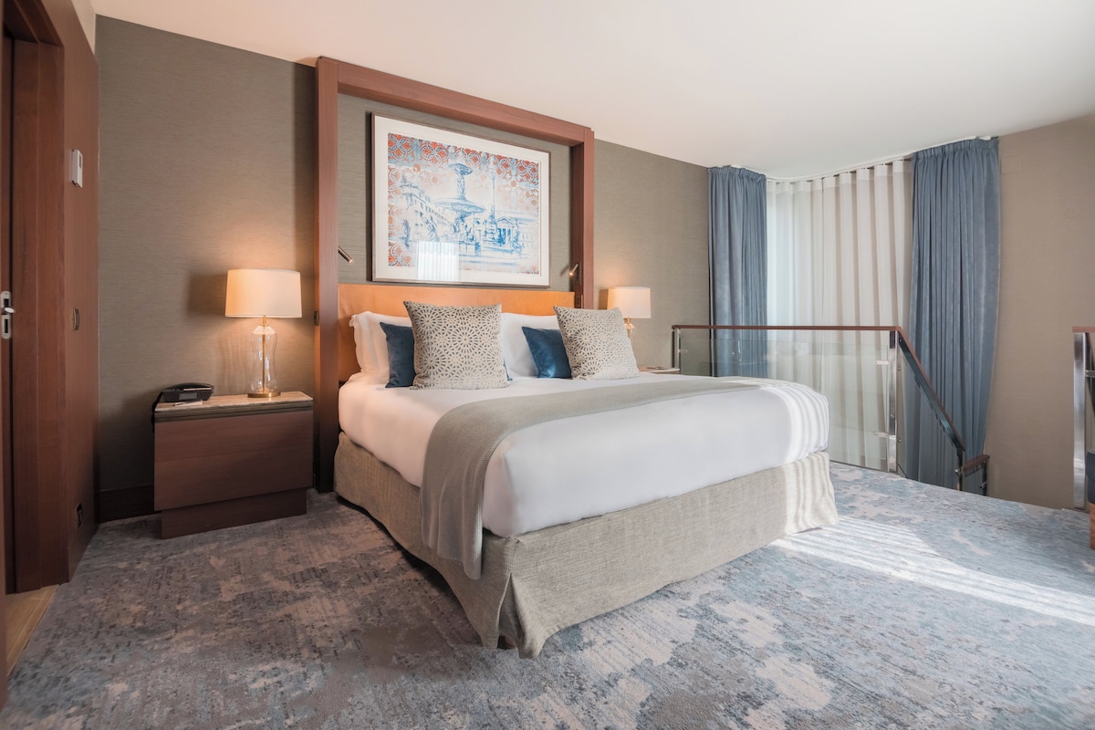 Habitación de hotel con una cama grande, dos mesitas de noche con lámparas, una obra de arte enmarcada sobre la cama, vistas al exterior a través de una ventana con cortinas, y una escalera a la derecha.