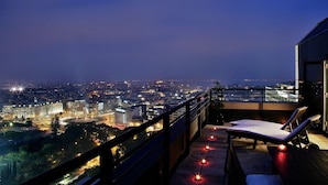 Een balkon met uitzicht op de stad 's nachts, met een zithoek en twee ligbedden en een balustrade.