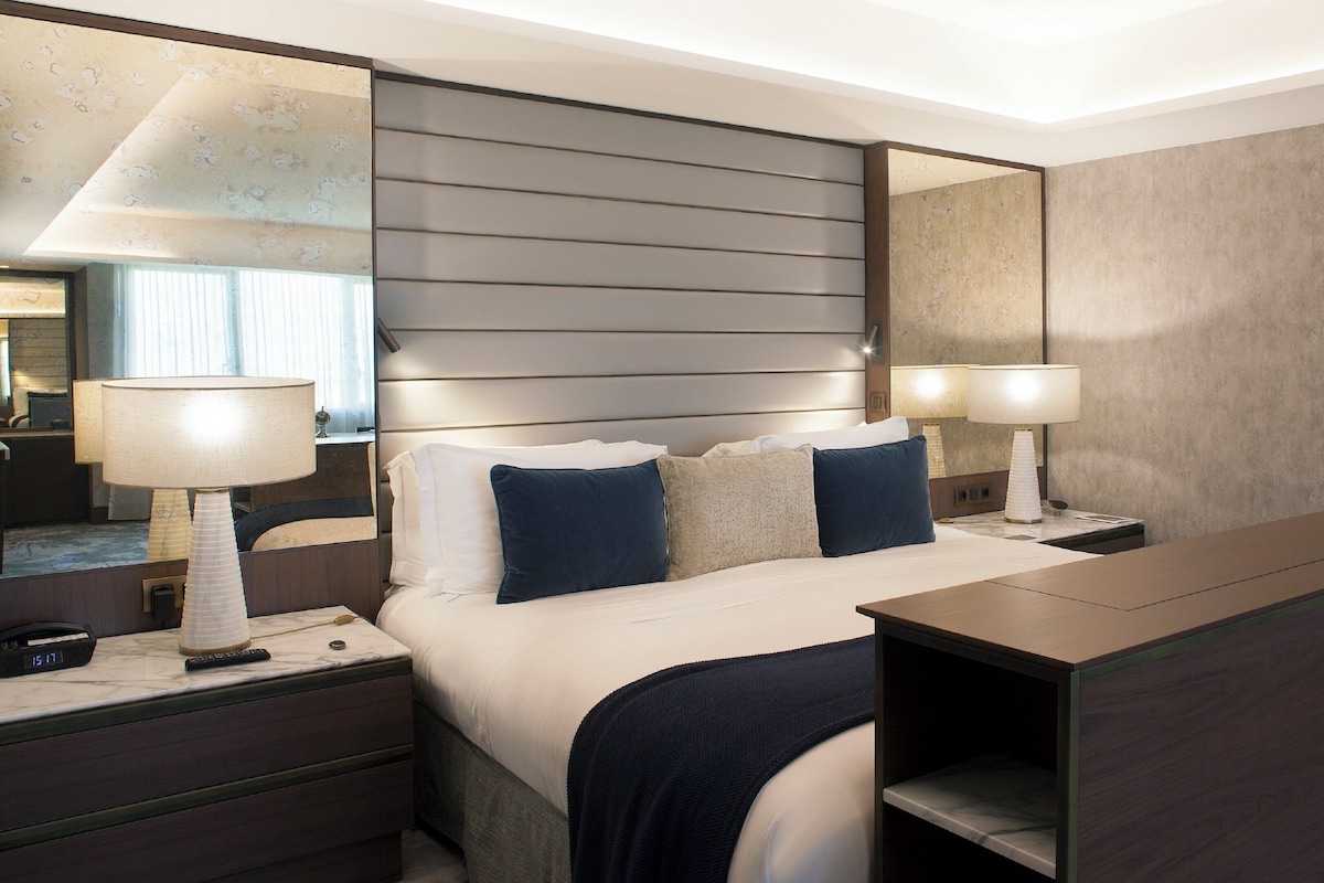 Una habitación de hotel con cama, mesitas de noche, lámparas, un espejo y un escritorio.