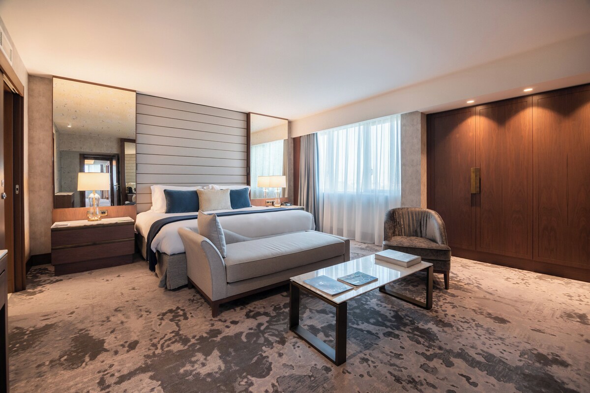 Habitación de hotel con cama, sofá, mesa de centro, sillón, mesita de noche, lámpara, espejo y armario.
