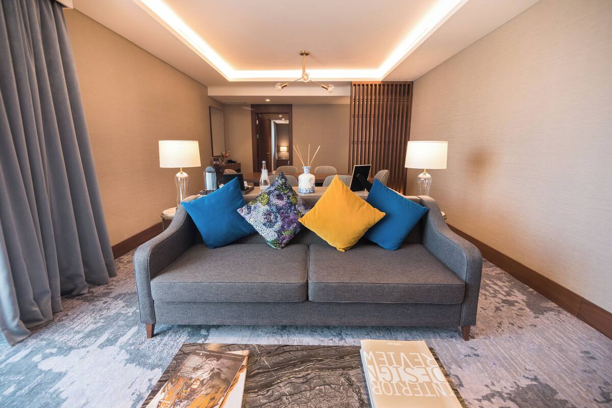 A modern hotel room with a grey sofa, two lamps, a coffee table, and a large window with curtains.