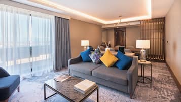 A modern hotel room with a grey sofa, a coffee table, and a view of the city through sheer curtains.
