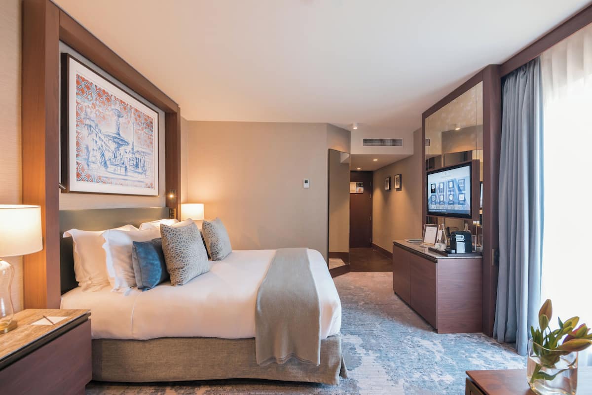 A hotel room with a large bed, bedside lamps, a desk with a flat-screen TV, and a framed artwork on the wall.