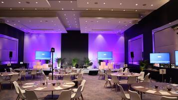 A conference room with multiple tables and chairs, equipped with projectors and screens, and a stage at the front.