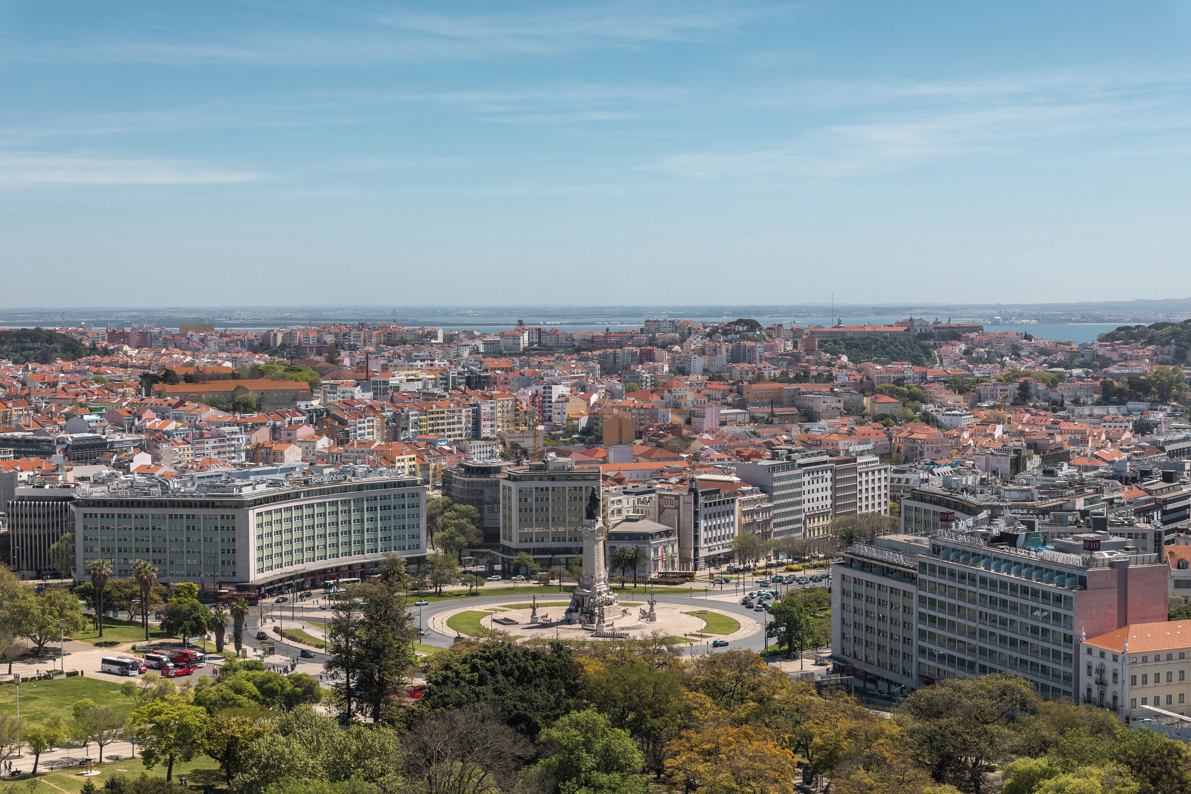 A cityscape with a prominent roundabout, various buildings, and a clear view of the sea.