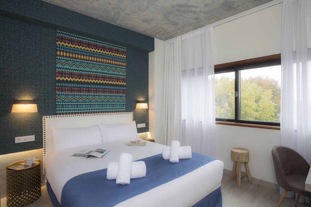 A hotel room with a large bed, two bedside lamps, a window with curtains, a small wooden stool, and a chair.
