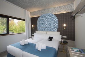 A hotel room with a large bed, two bedside tables, a window with curtains, a patterned wall, and a view of trees outside.