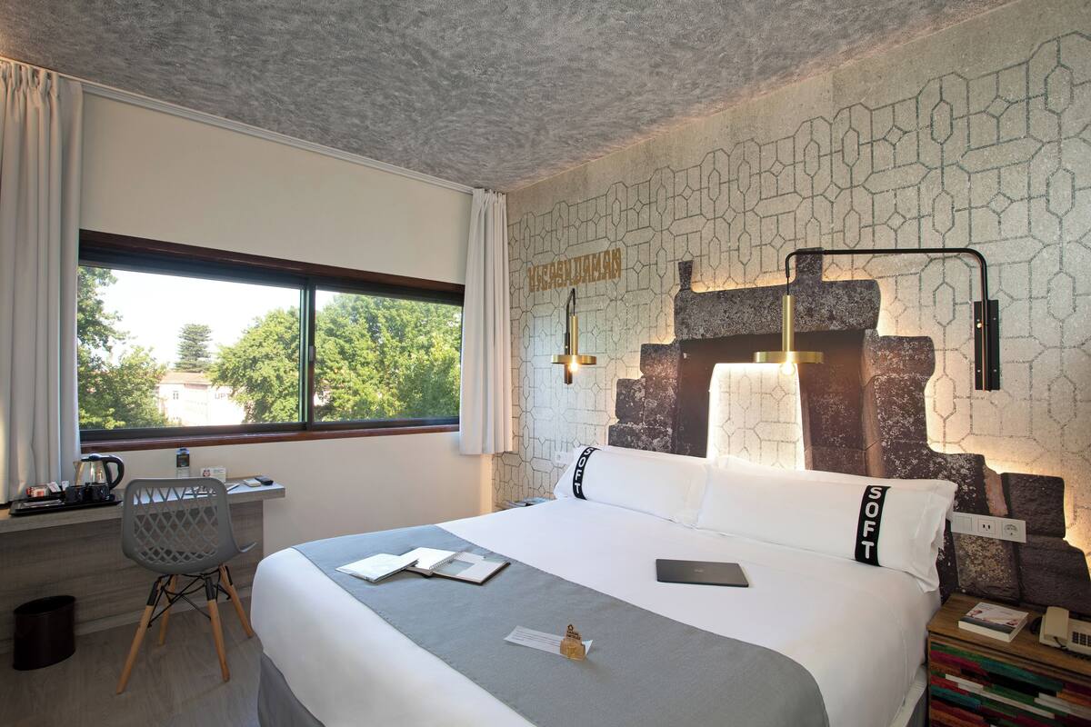A hotel room with a large bed, a desk with a chair, a window with curtains, and a unique wall design.