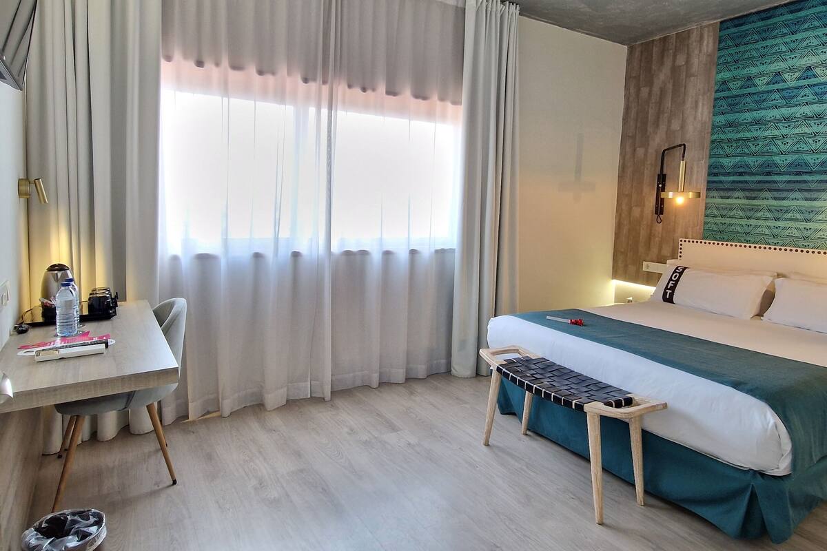 A hotel room with a bed, a desk, a chair, a bench, and a large window with curtains.
