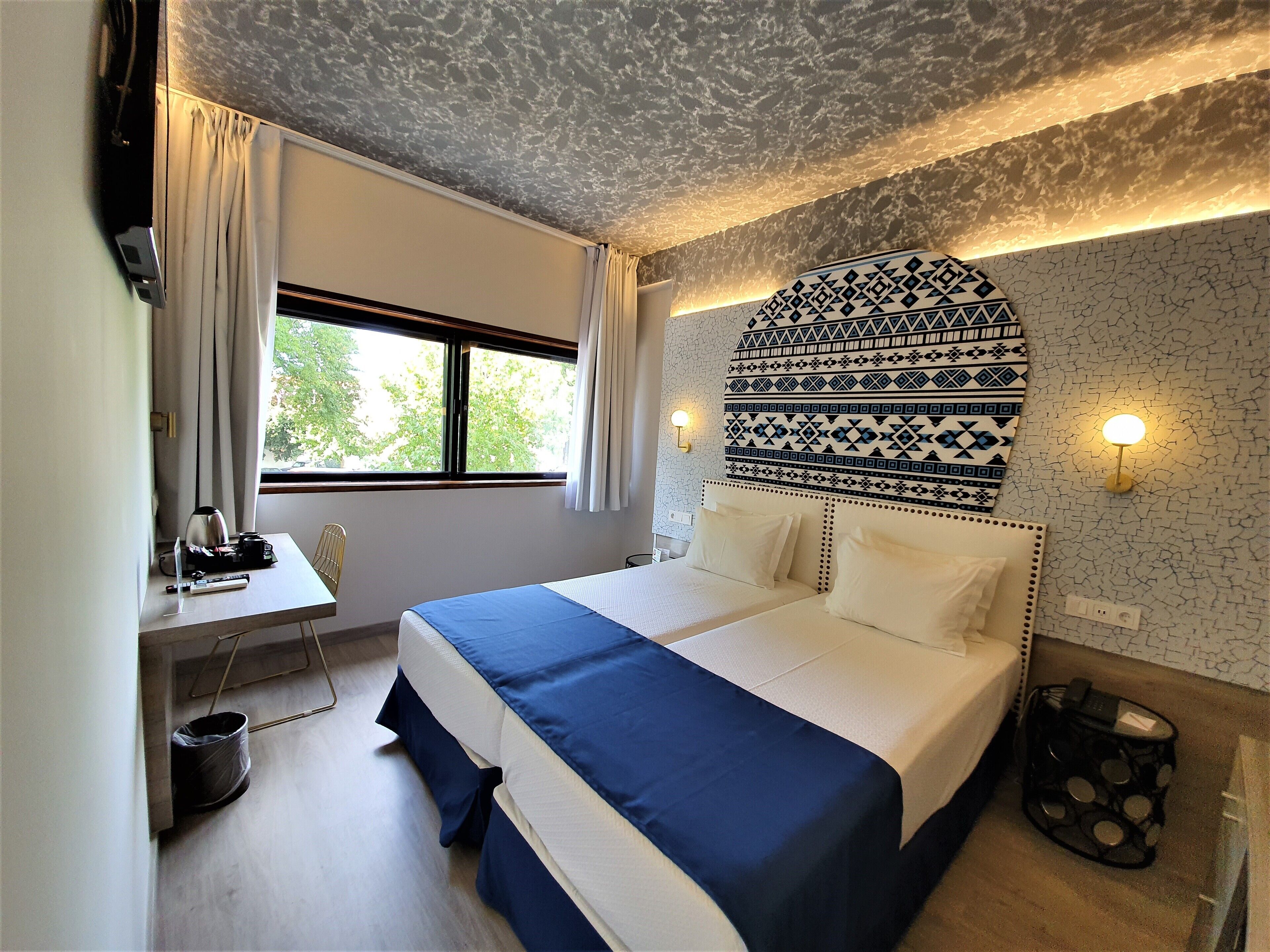 Hotellihuone, jossa on suuri sänky, työpöytä tuolilla, ikkuna verhoilla ja kuvioitu seinä.