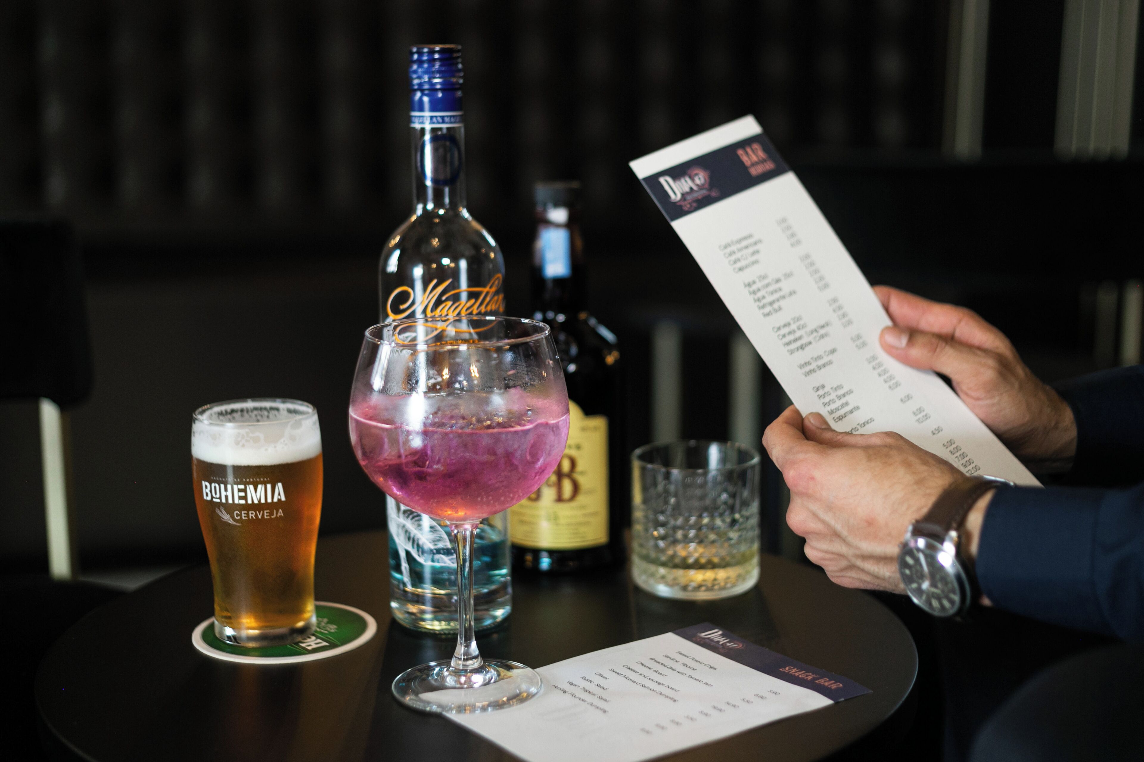 A table with a beer glass labeled "BOHEMIA CERVEJA," a cocktail glass with a purple drink, a bottle of liquor, and a menu.