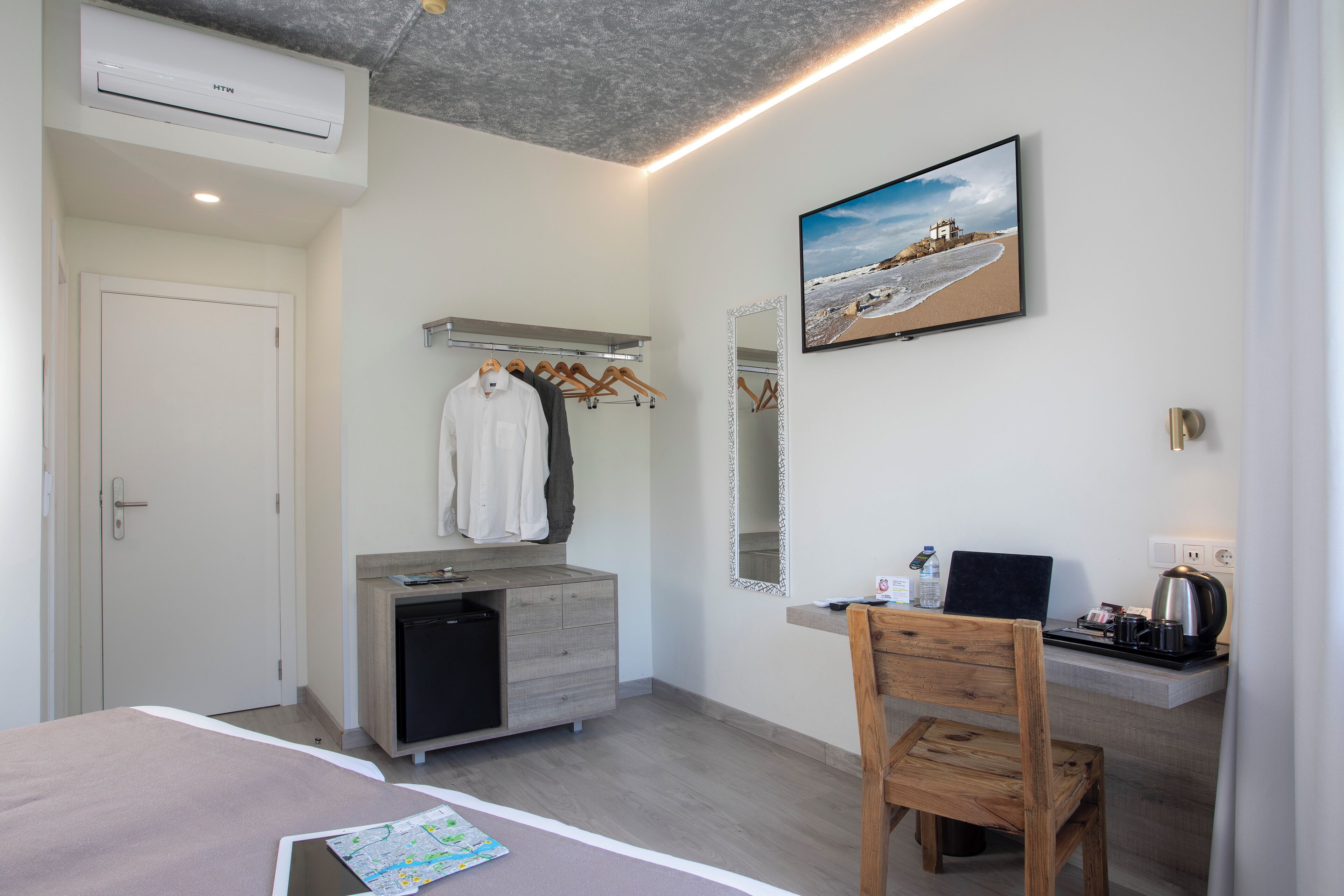 A modern hotel room with a bed, a desk with a chair, a small kitchenette, a mirror, and a painting on the wall.