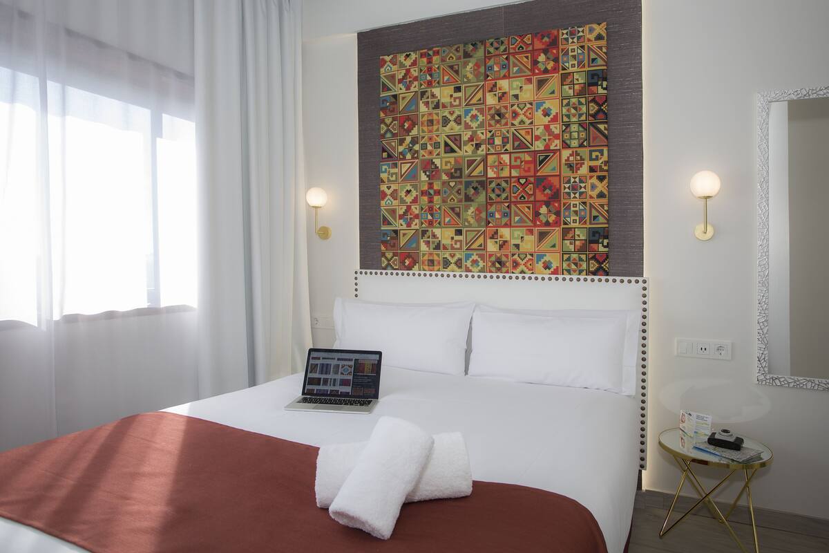 A hotel room with a bed, a laptop, a bedside table with a lamp, and a colorful tapestry on the wall.