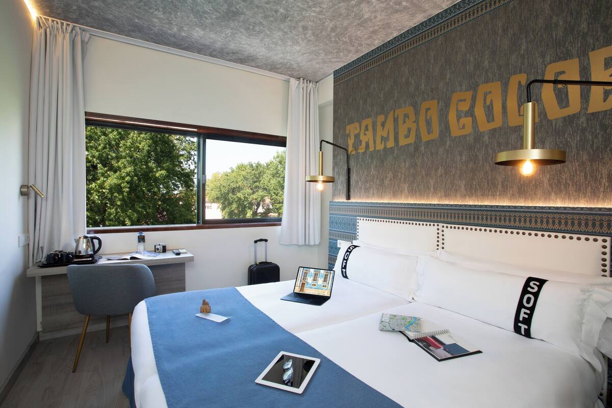 A hotel room with a bed, a desk, a laptop, and a view of trees through the window.
